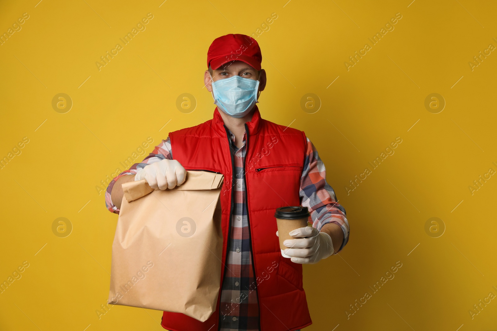 Photo of Courier in protective mask and gloves holding order on yellow background. Food delivery service during coronavirus quarantine