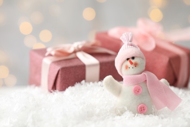 Photo of Snowman toy and Christmas gift boxes on snow against blurred festive lights. Space for text