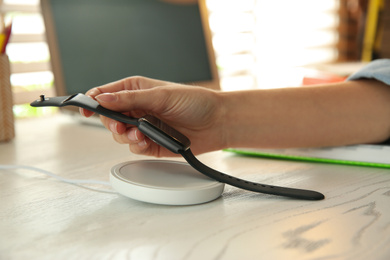 Photo of Woman putting smartwatch onto wireless charger at white wooden table, closeup. Modern workplace accessory