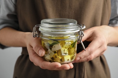 Photo of Woman holding jar with pickled feta cheese on grey background, closeup