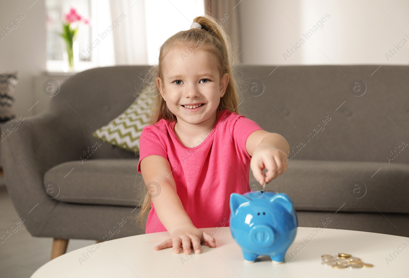 Photo of Cute girl putting coin into piggy bank at table in living room. Saving money