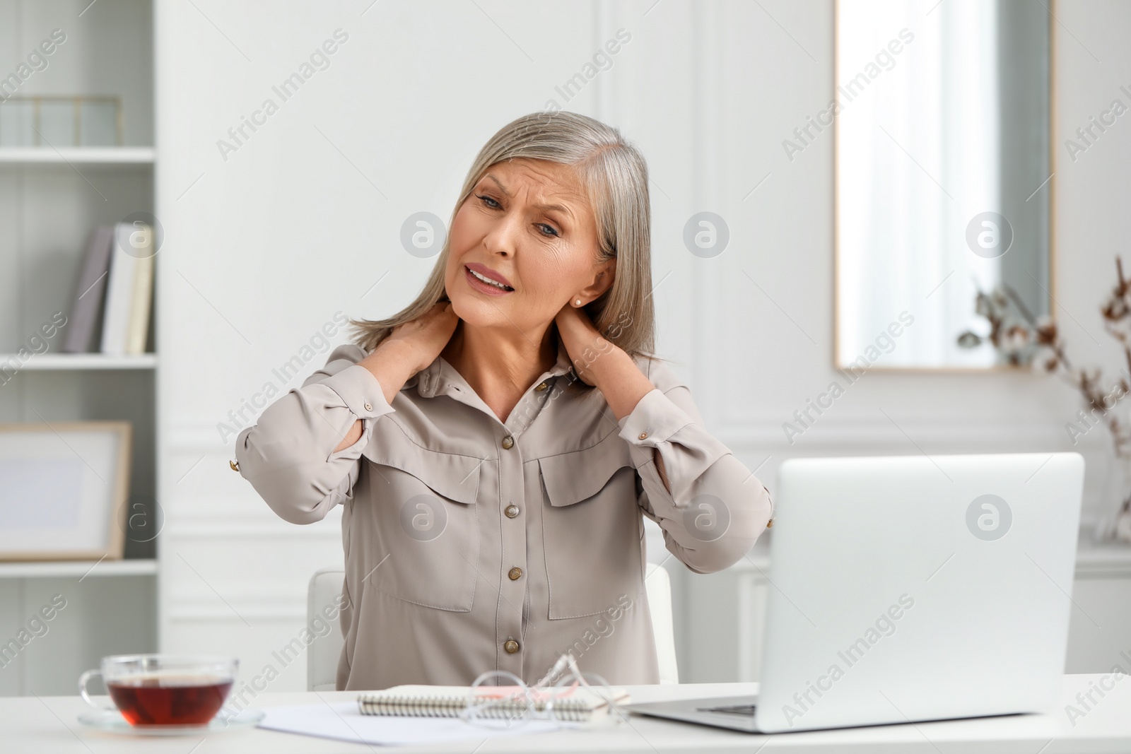 Photo of Woman suffering from neck pain at workplace in room