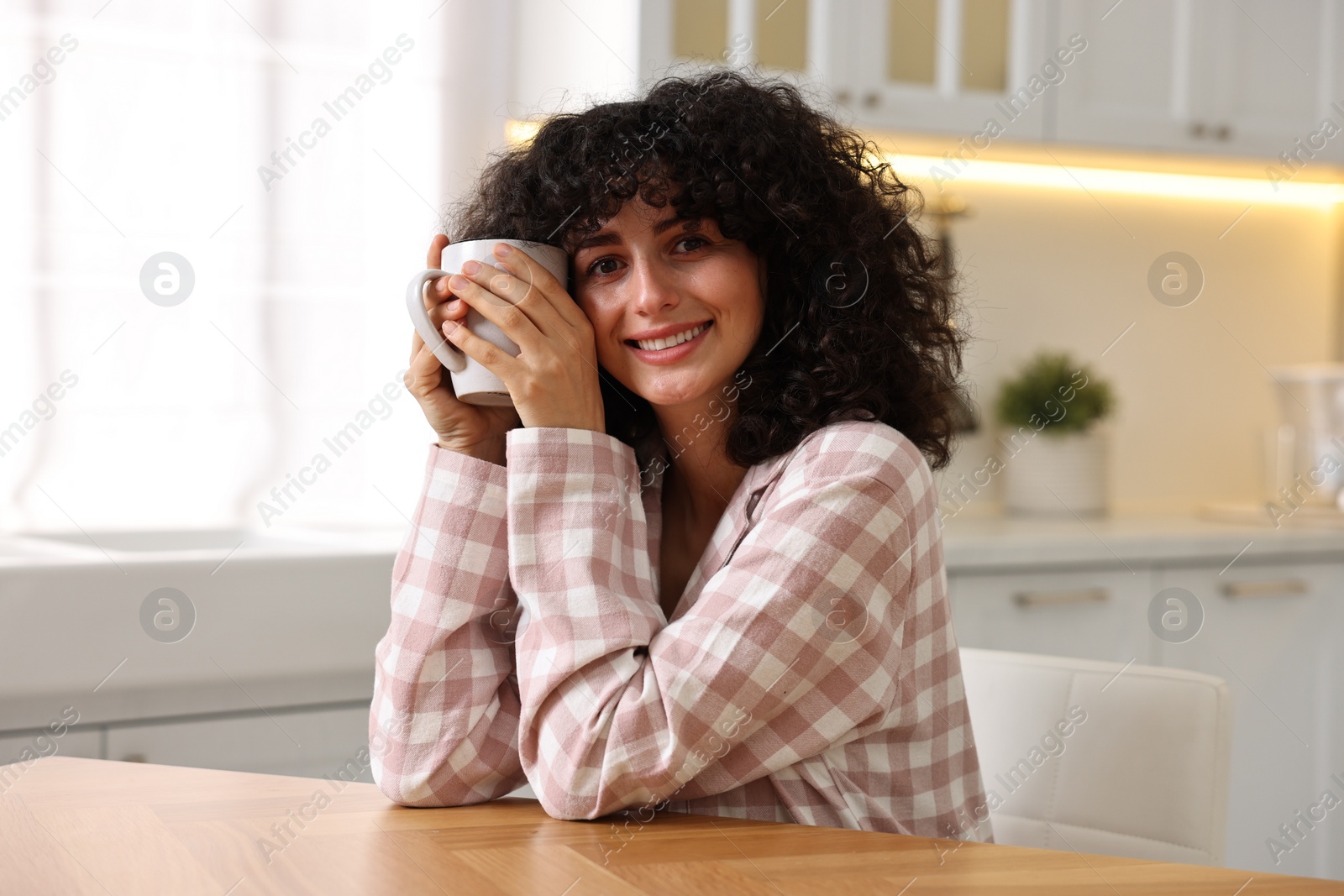 Photo of Beautiful young woman in stylish pyjama with cup of drink at wooden table in kitchen