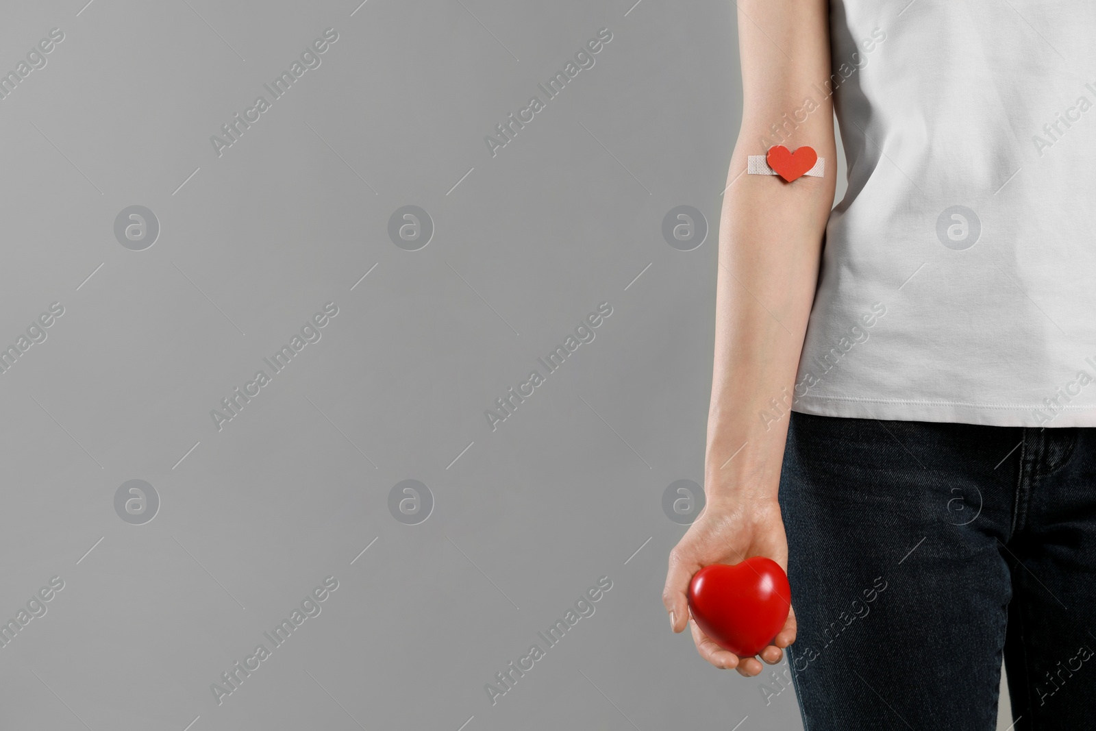 Photo of Blood donation concept. Woman with adhesive plaster on arm holding red heart against grey background, closeup. Space for text