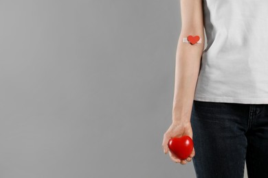 Blood donation concept. Woman with adhesive plaster on arm holding red heart against grey background, closeup. Space for text