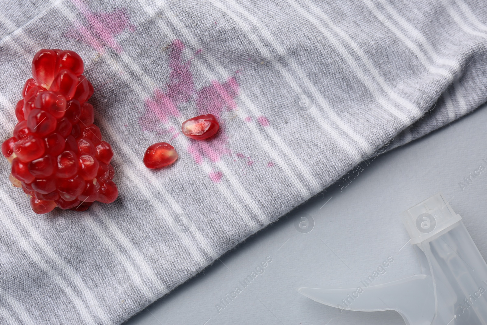 Photo of Striped shirt with fruit juicy stains, detergent and pomegranate seeds on light grey background, above view