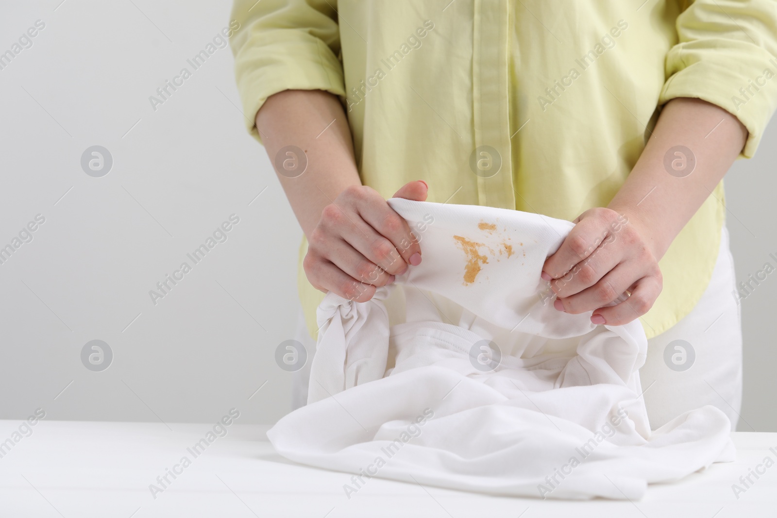 Photo of Woman holding shirt with stain at white table against light grey background, closeup