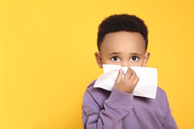 Photo of African-American boy blowing nose in tissue on yellow background, space for text. Cold symptoms