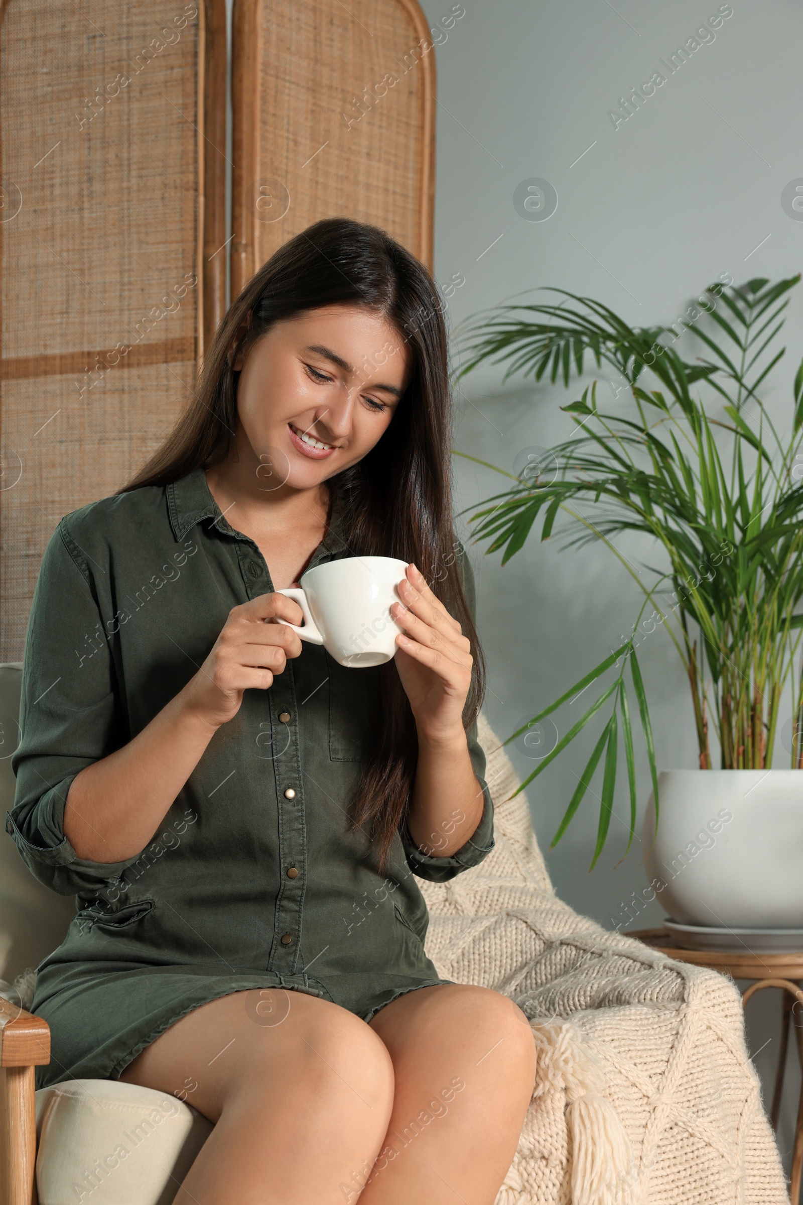 Photo of Happy young woman with cup of coffee sitting in armchair at home