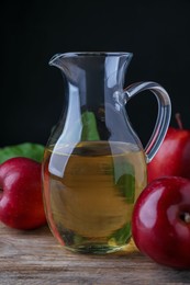 Photo of Jug of tasty juice and fresh red apples on wooden table against black background