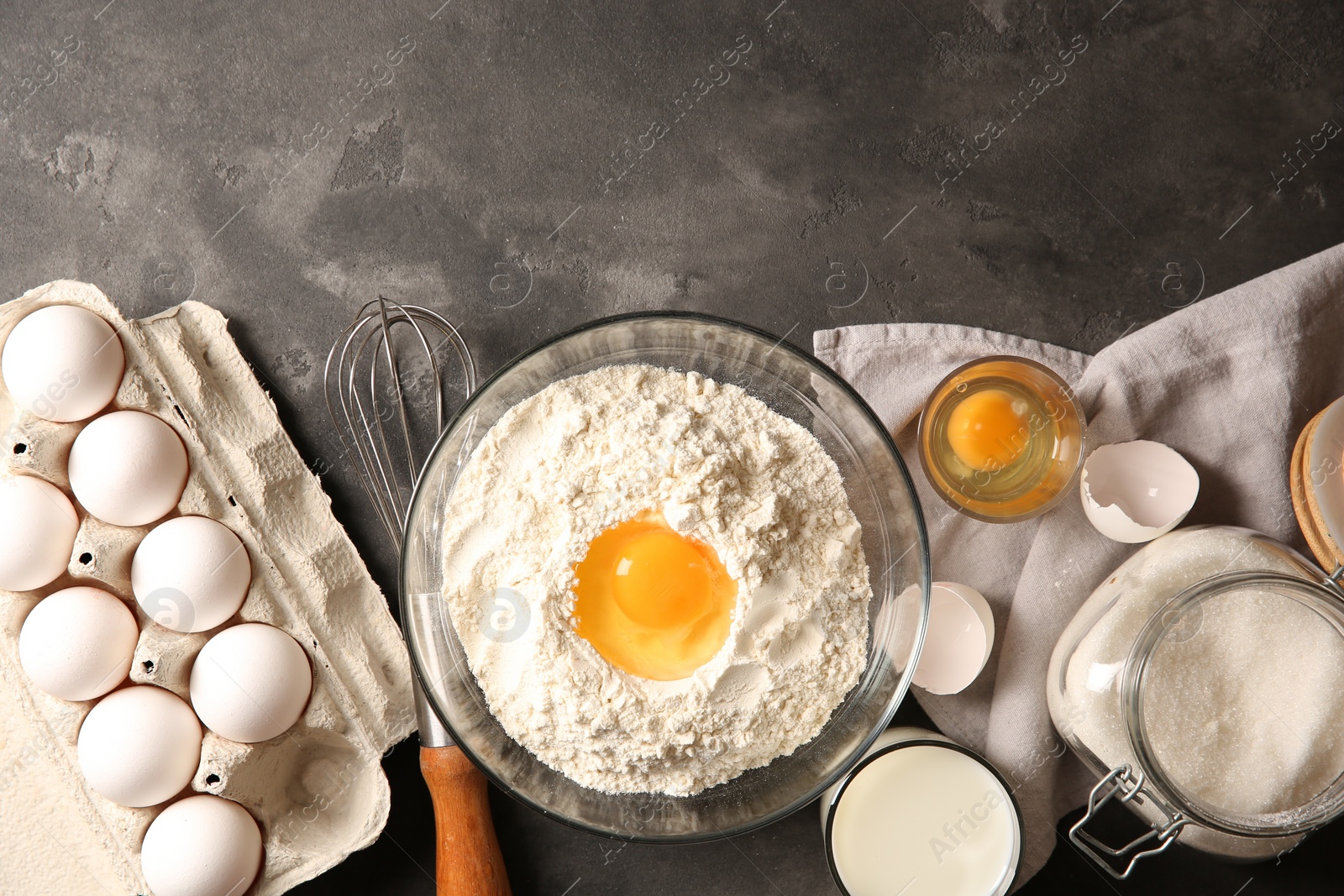 Photo of Making dough. Flour with egg yolk in bowl and other products on grey textured table, flat lay