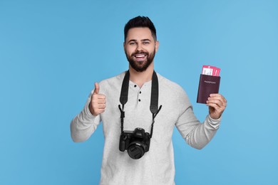 Photo of Happy man with passport, tickets and camera showing thumb up on light blue background