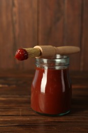 Tasty barbecue sauce in glass jar and brush on wooden table, closeup