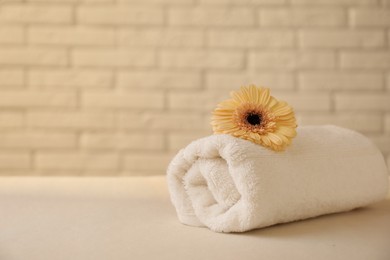 Rolled terry towel and flower on white table near brick wall indoors, closeup. Space for text