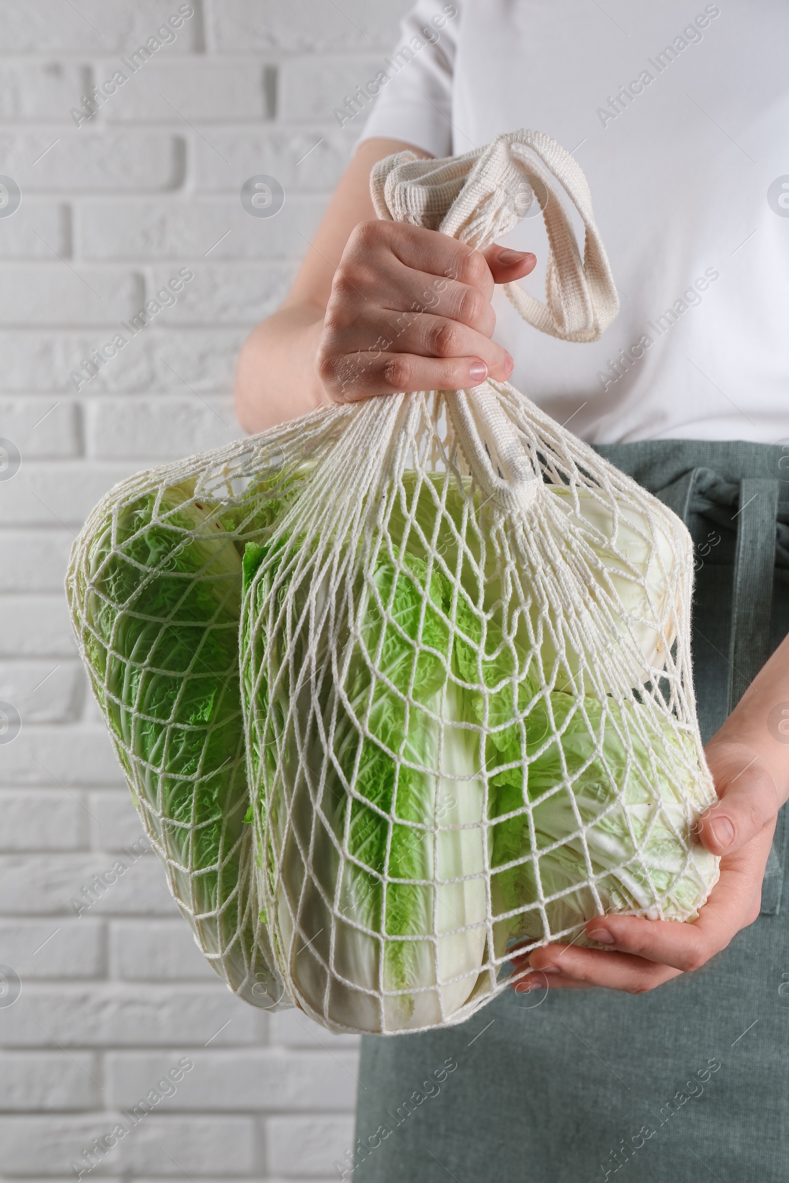 Photo of Woman holding string bag with fresh Chinese cabbages near white brick wall, closeup