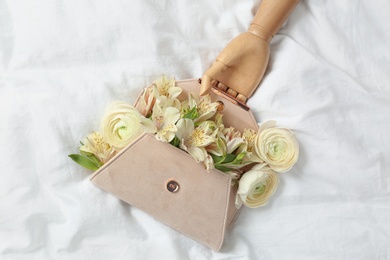 Photo of Stylish clutch with spring flowers and wooden mannequin hand on crumpled sheet, flat lay