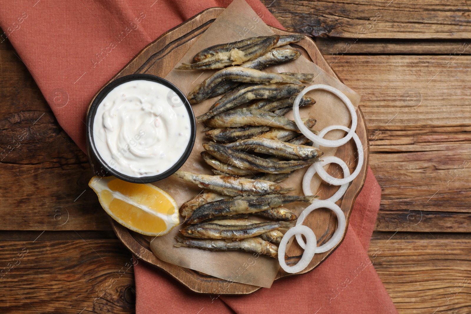 Photo of Delicious fried anchovies with onion rings, sauce and slice of lemon served on wooden table, top view. Space for text