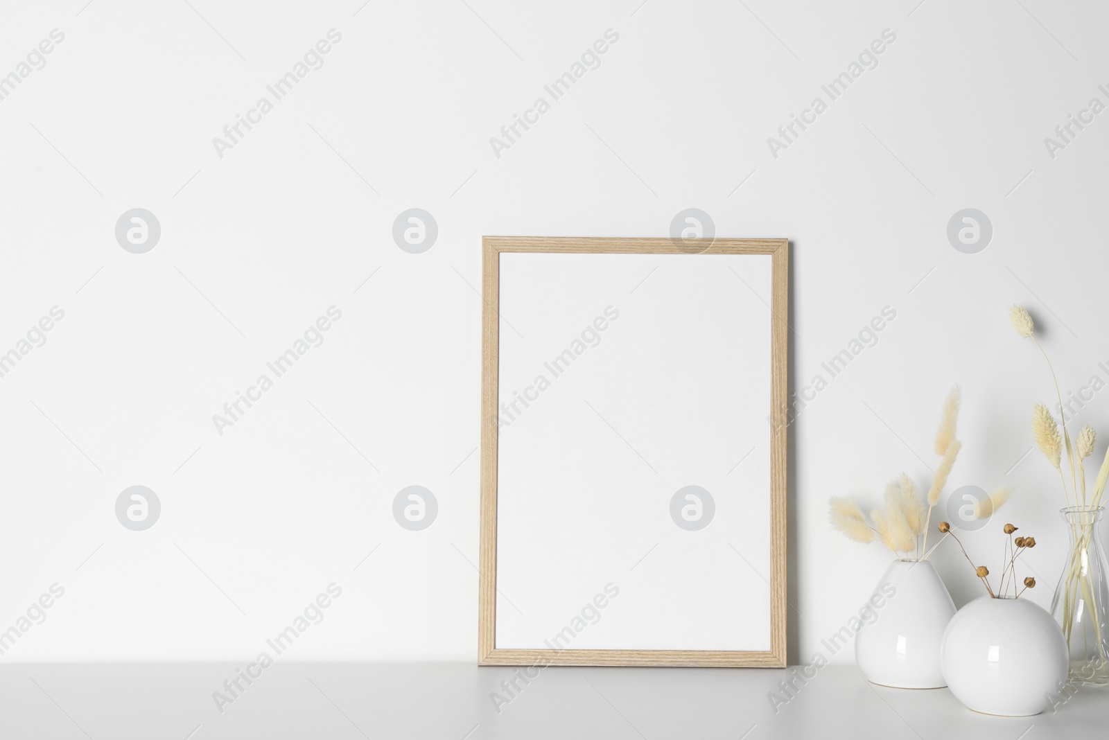 Photo of Empty photo frame and vases with dry decorative spikes on white table. Mockup for design