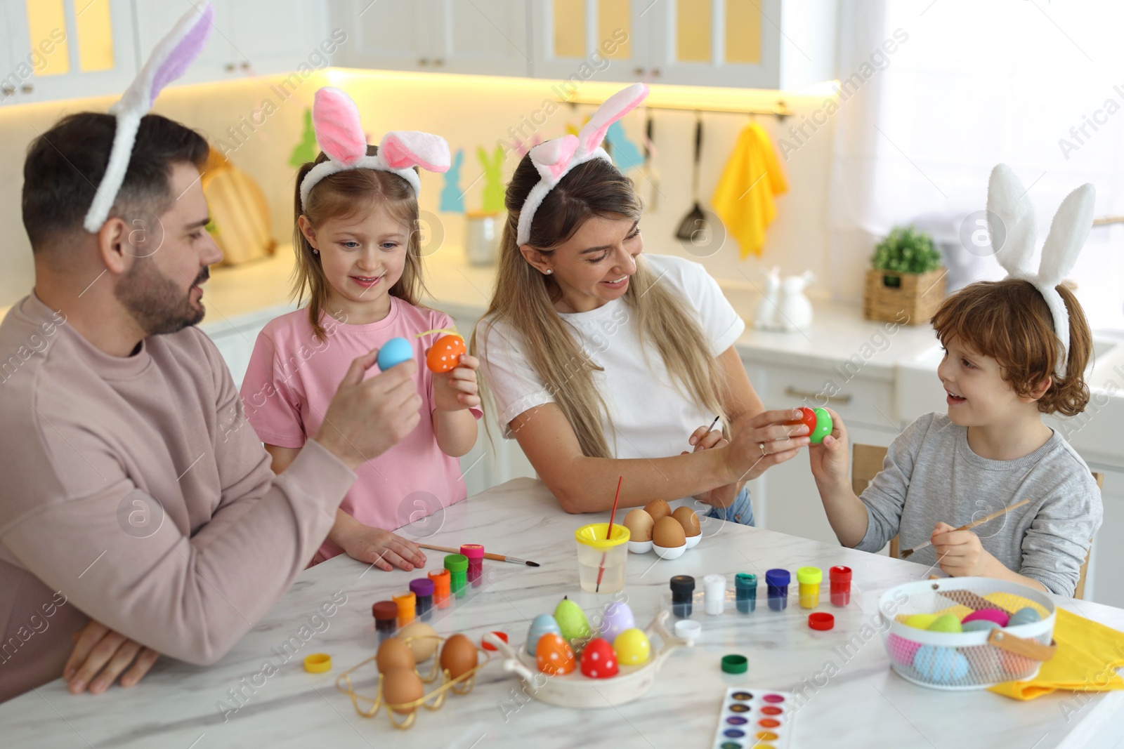 Photo of Easter celebration. Happy family with bunny ears having fun while painting eggs at white marble table in kitchen