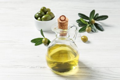 Photo of Glass jug of oil, ripe olives and green leaves on white wooden table