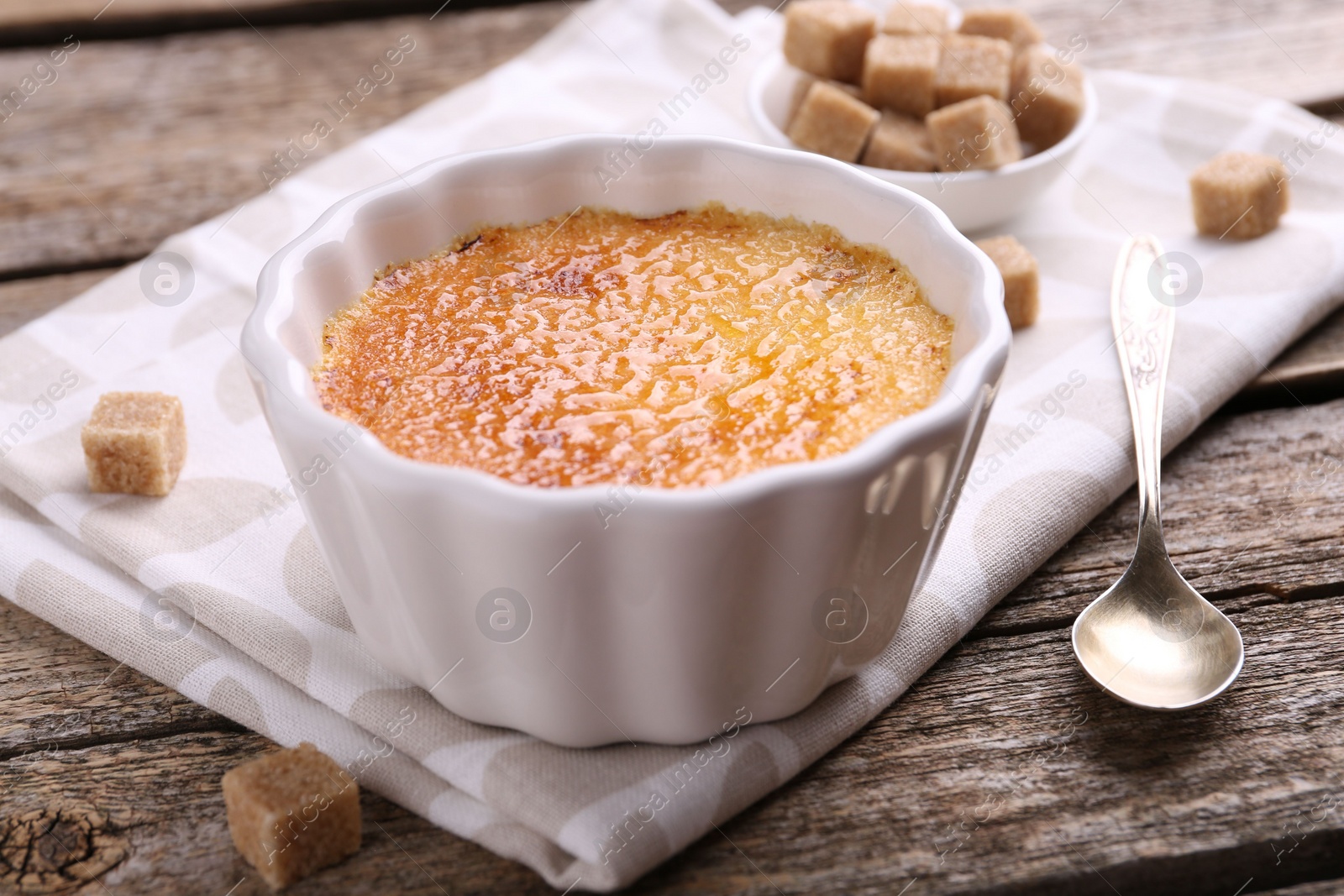 Photo of Delicious creme brulee in bowl, sugar cubes and spoon on wooden table, closeup