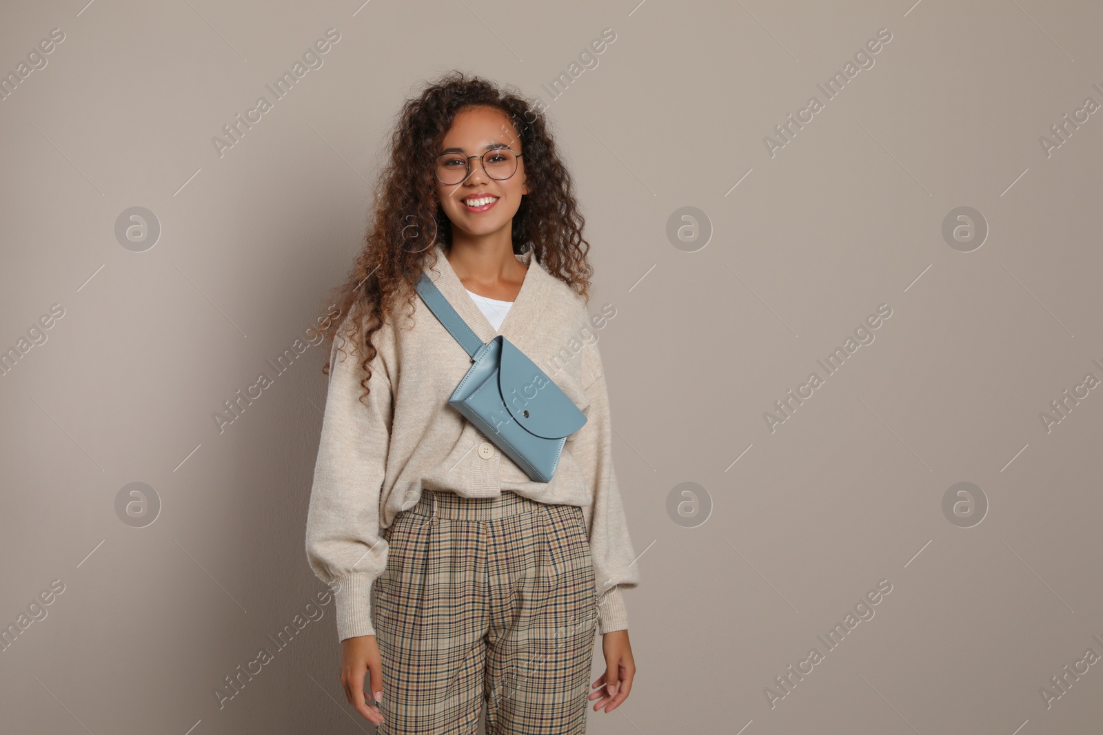 Photo of Beautiful African American woman with stylish waist bag on beige background, space for text