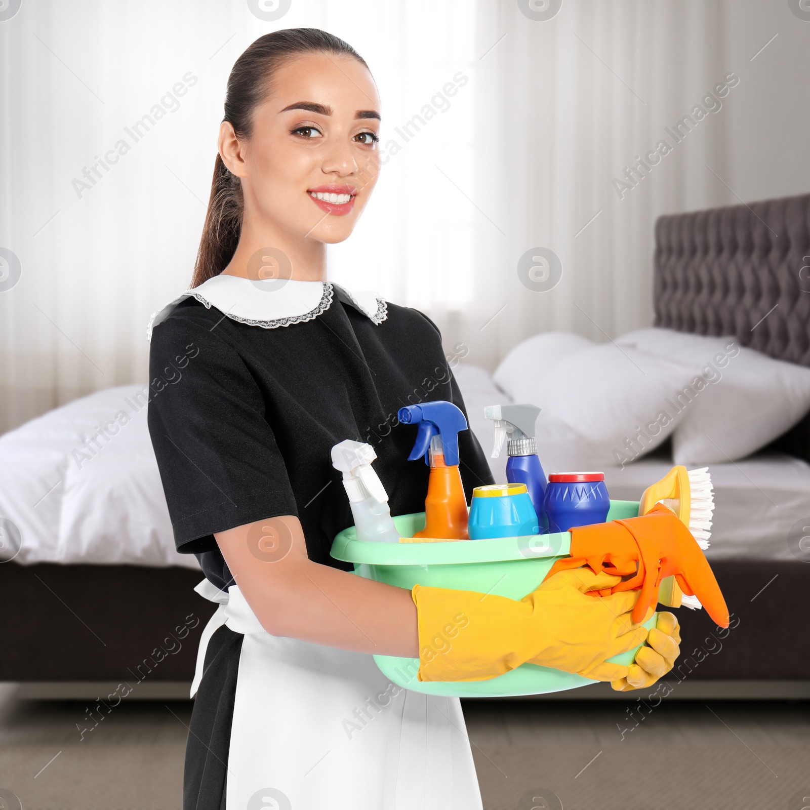 Image of Beautiful chambermaid with cleaning supplies near bed in hotel room