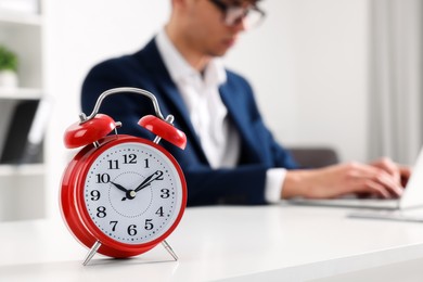 Photo of Red alarm clock and man working at table in office, closeup with space for text. Deadline concept