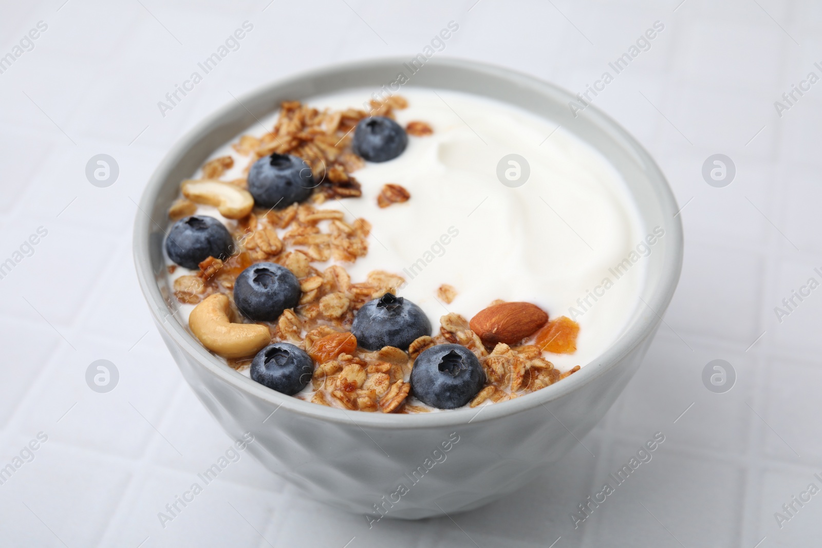 Photo of Bowl with yogurt, blueberries and granola on white tiled table, closeup