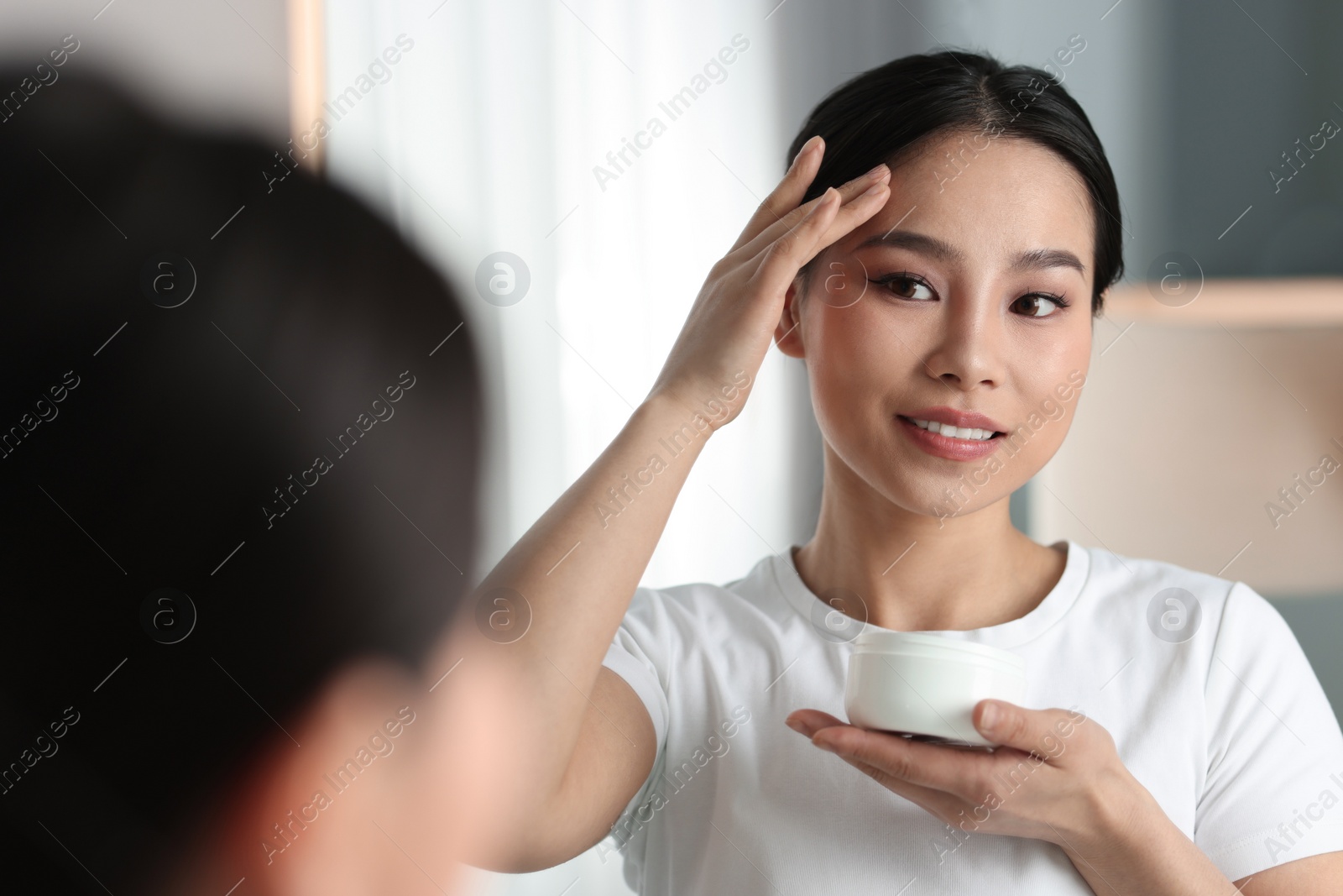 Photo of Happy woman applying face cream near mirror at home