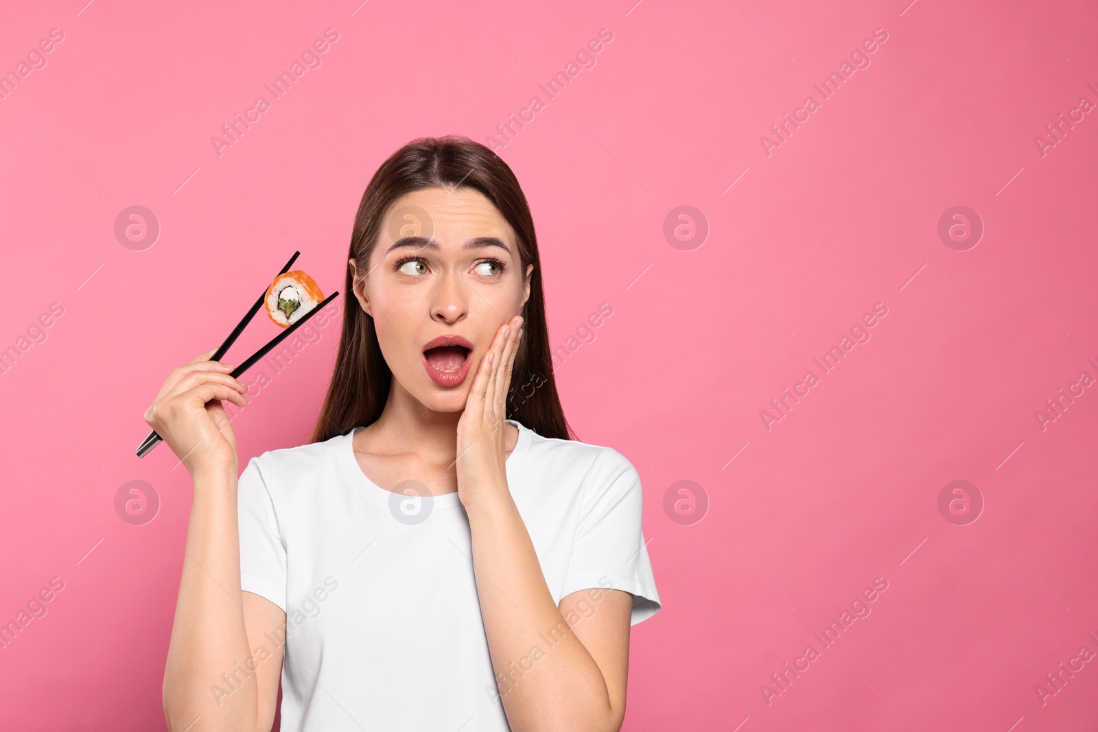 Photo of Emotional young woman holding sushi roll with chopsticks on pink background. Space for text