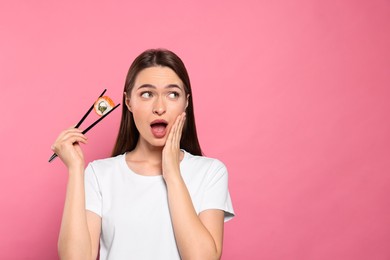 Emotional young woman holding sushi roll with chopsticks on pink background. Space for text