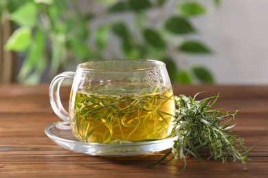 Photo of Cup of homemade herbal tea and fresh tarragon leaves on wooden table, closeup
