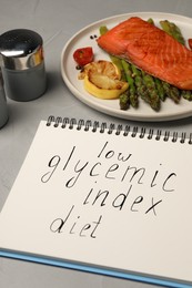 Photo of Notebook with words Low Glycemic Index Diet and plate of tasty grilled salmon on grey table