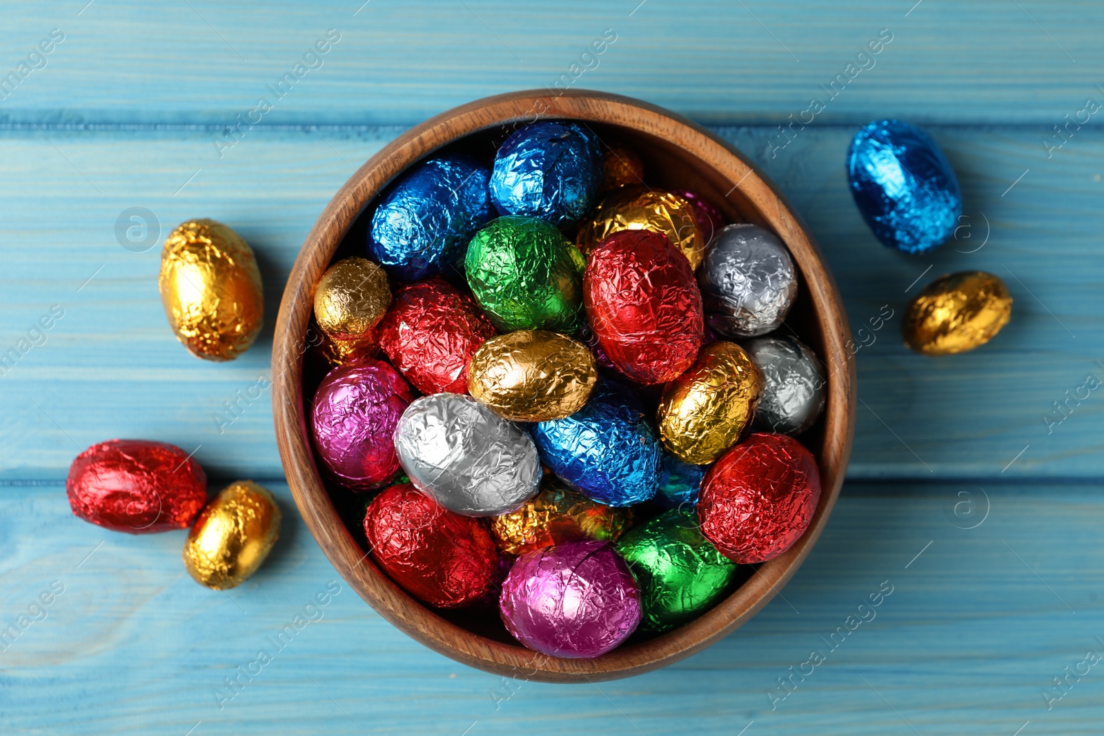 Photo of Bowl with chocolate eggs wrapped in colorful foil on light blue wooden table, flat lay