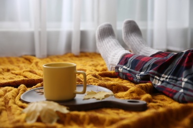 Woman relaxing with cup of hot winter drink on knitted plaid indoors, closeup. Cozy season