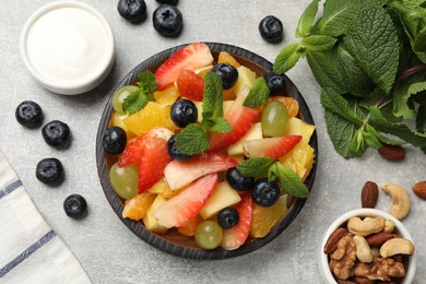 Delicious fruit salad in bowl, berries, nuts and fresh mint on grey table, flat lay