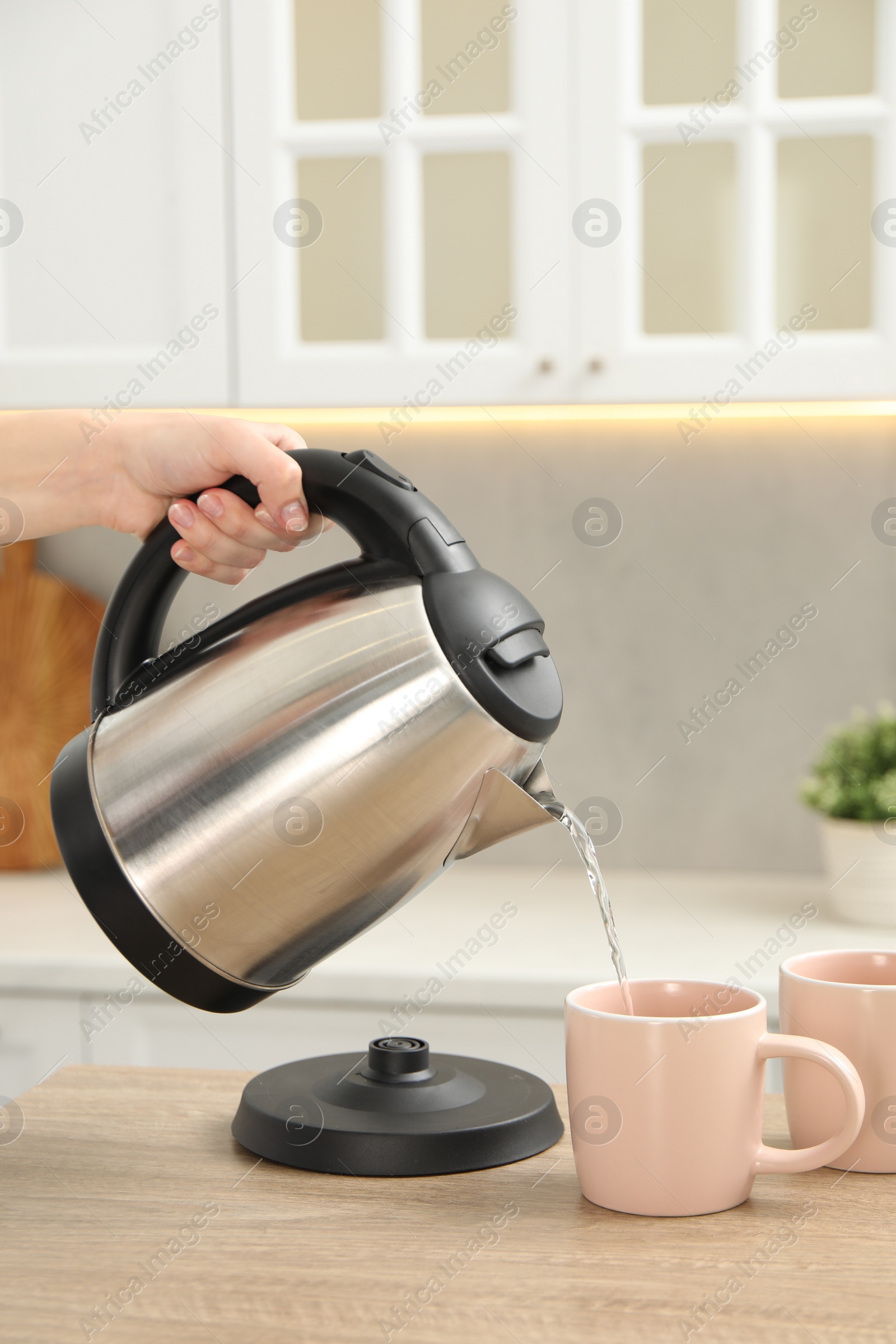 Photo of Woman pouring hot water from electric kettle into cup in kitchen, closeup