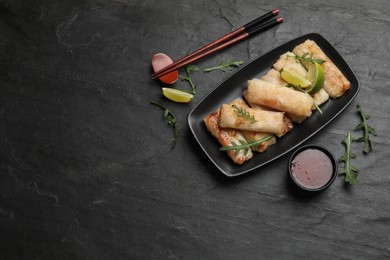 Photo of Tasty fried spring rolls, arugula, lime and sauce on dark textured table, flat lay. Space for text