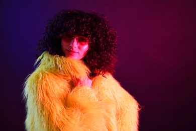 Photo of Beautiful young woman in yellow fur coat and sunglasses on color background in neon lights. Space for text