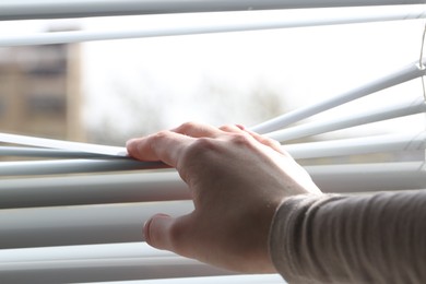 Woman separating slats of white blinds indoors, closeup