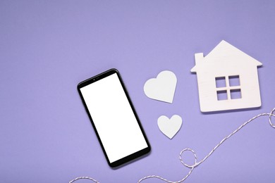 Photo of Long-distance relationship concept. Smartphone, white house, paper hearts and decorative cord on violet background, flat lay