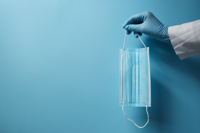 Photo of Doctor in latex gloves holding disposable face mask on light blue background, closeup with space for text. Protective measures during coronavirus quarantine