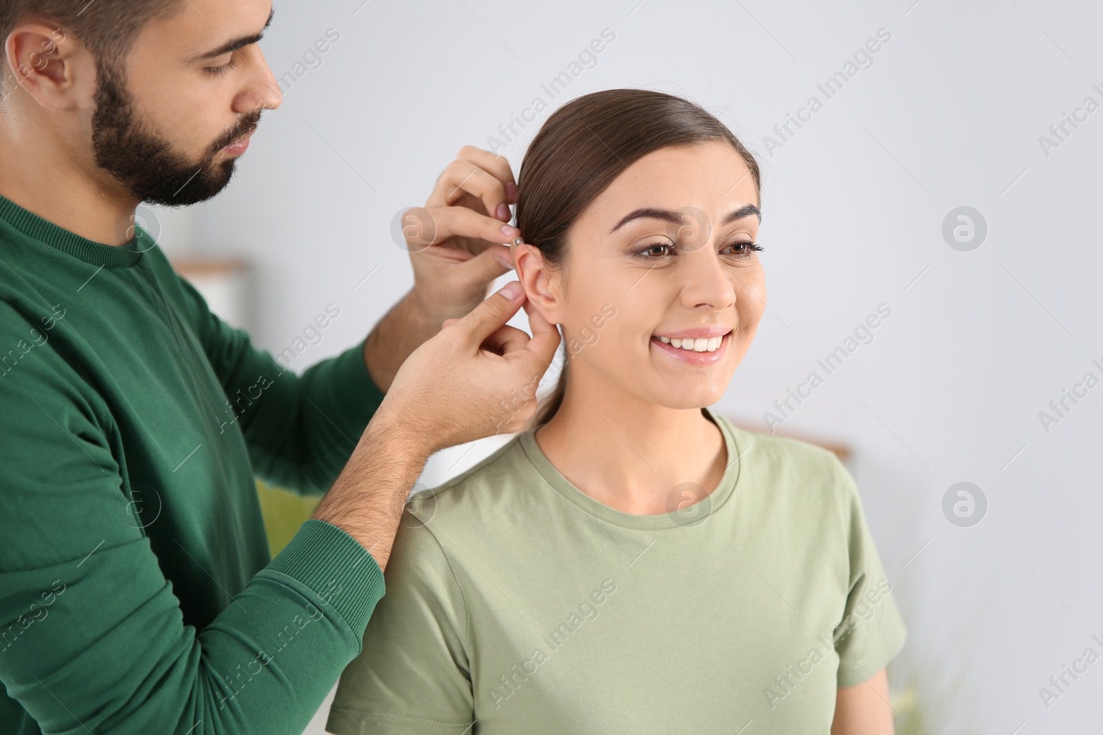 Photo of Young man putting hearing aid in woman's ear indoors