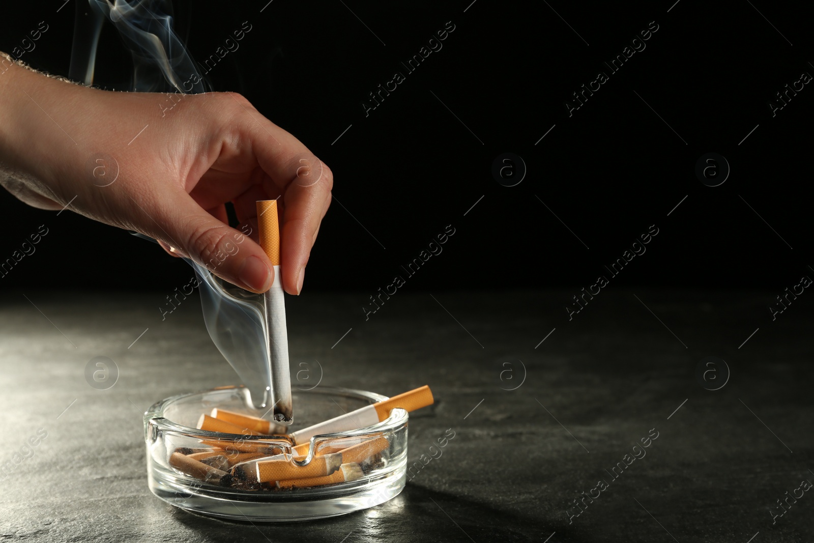 Photo of Woman extinguishing cigarette in glass ashtray
at grey table against black background, closeup