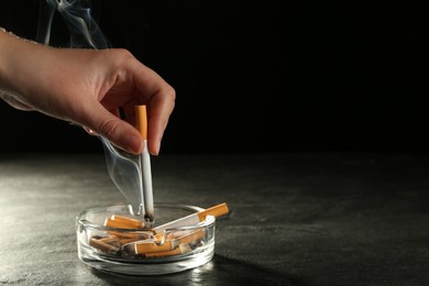 Woman extinguishing cigarette in glass ashtray
at grey table against black background, closeup