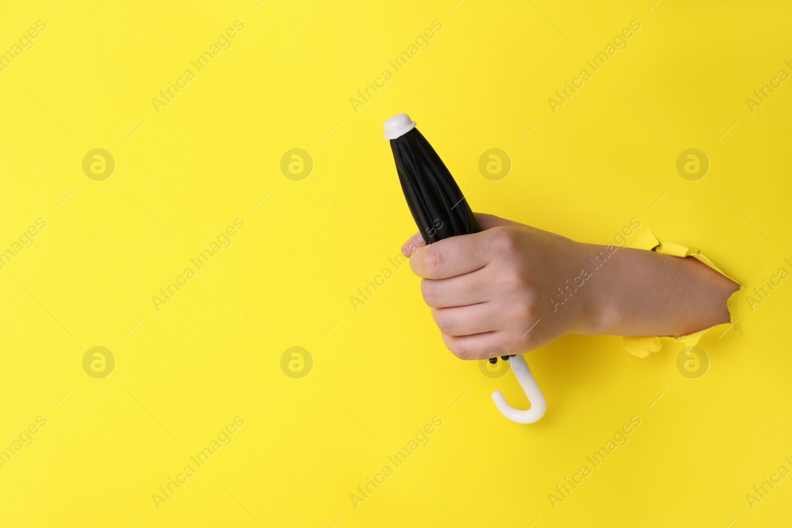 Photo of Woman holding closed small black umbrella through hole in yellow paper, closeup. Space for text