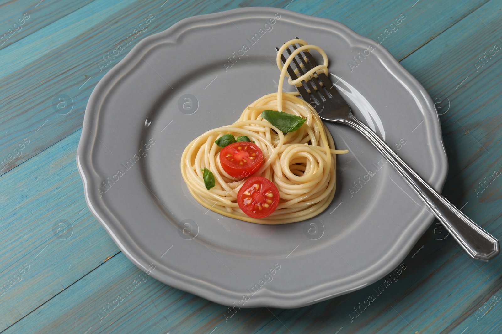 Photo of Heart made of tasty spaghetti, fork, tomato and basil on light blue wooden table