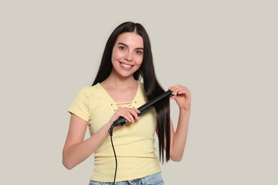 Beautiful happy woman using hair iron on light grey background