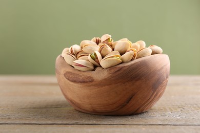 Photo of Tasty pistachios in bowl on wooden table against olive background, closeup